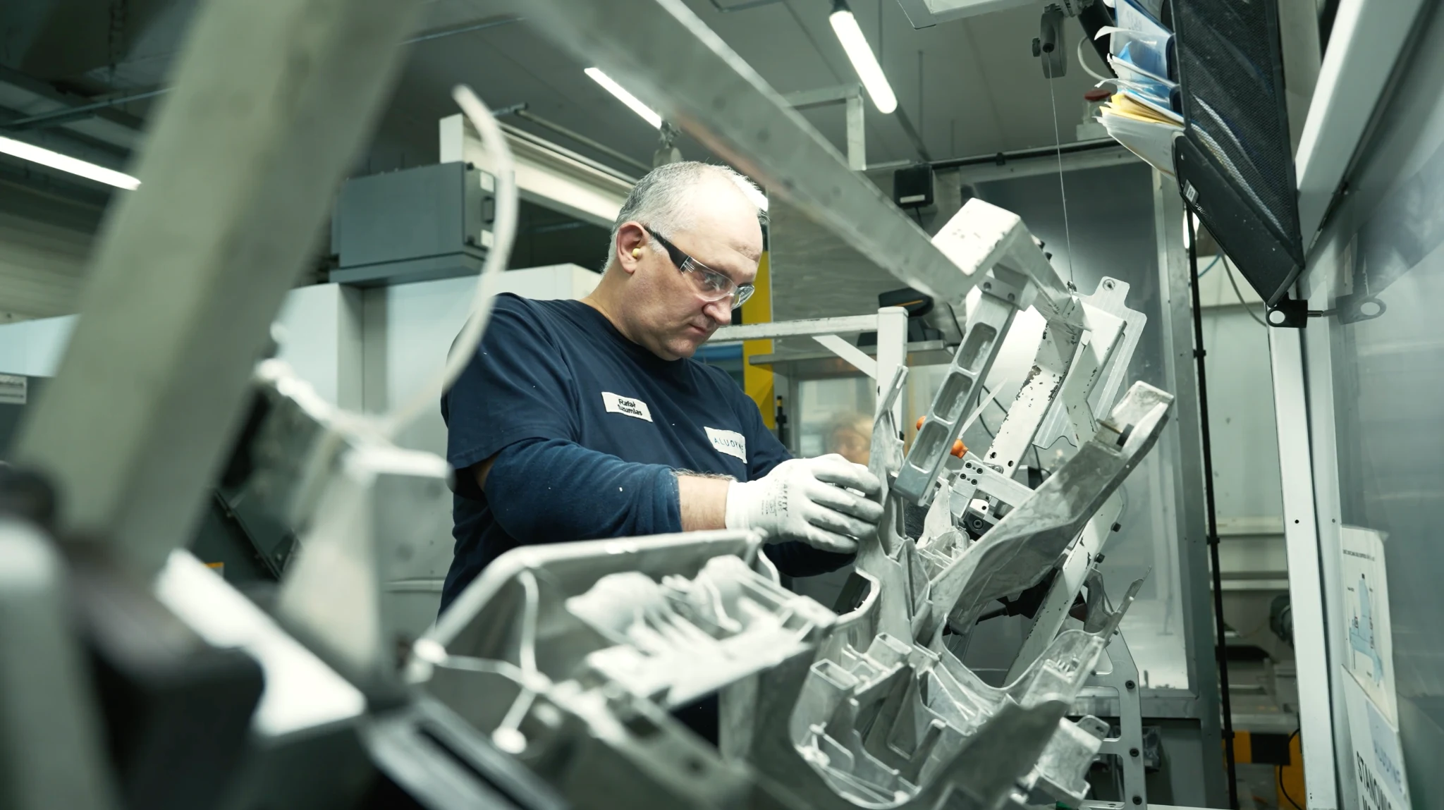 A factory worker in safety glasses and gloves inspecting cast aluminum components on a production line